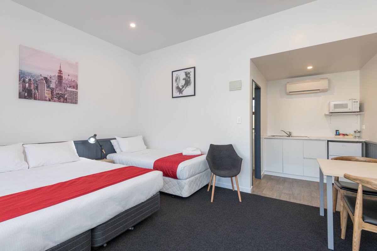 A hotel room with two beds, a desk, a chair, a kitchenette, and a cityscape painting.