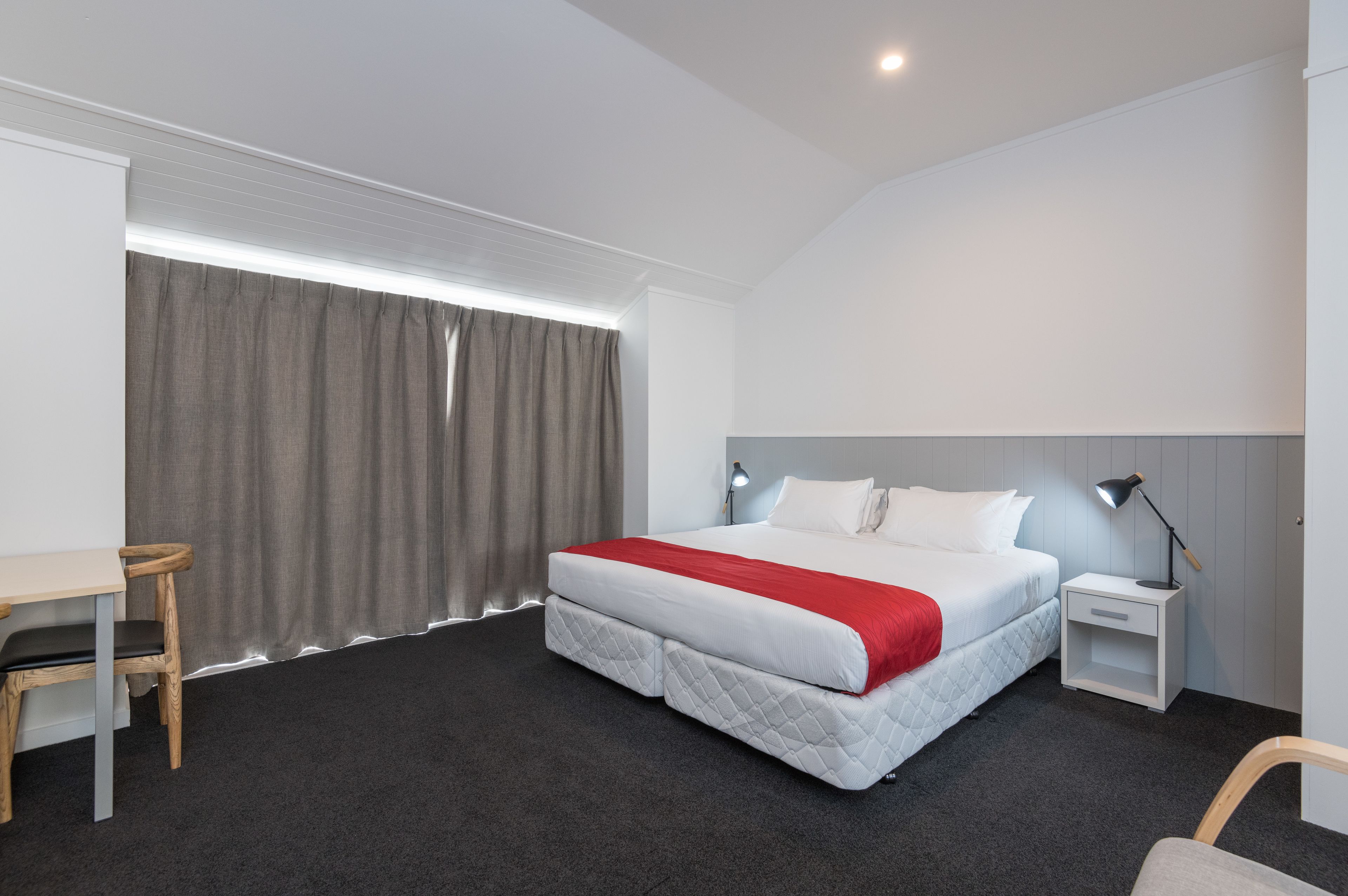 A hotel room with a bed, bedside table, lamp, and a desk with a chair.