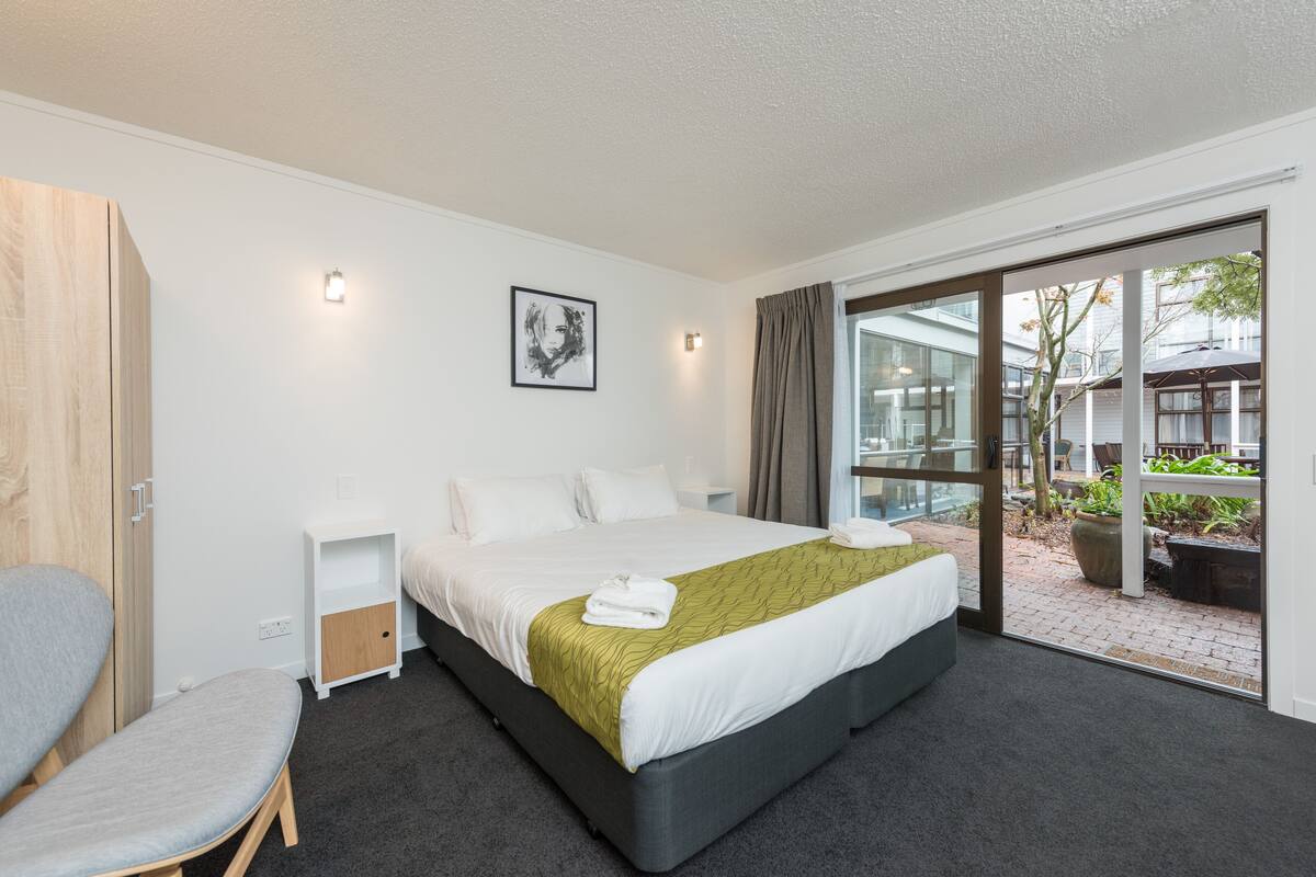A modern hotel room with a large bed, a small bedside table, a chair, and a view of the outdoor area through a glass door.