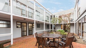 A courtyard with a round wooden table and chairs, surrounded by a brick floor and white railings.