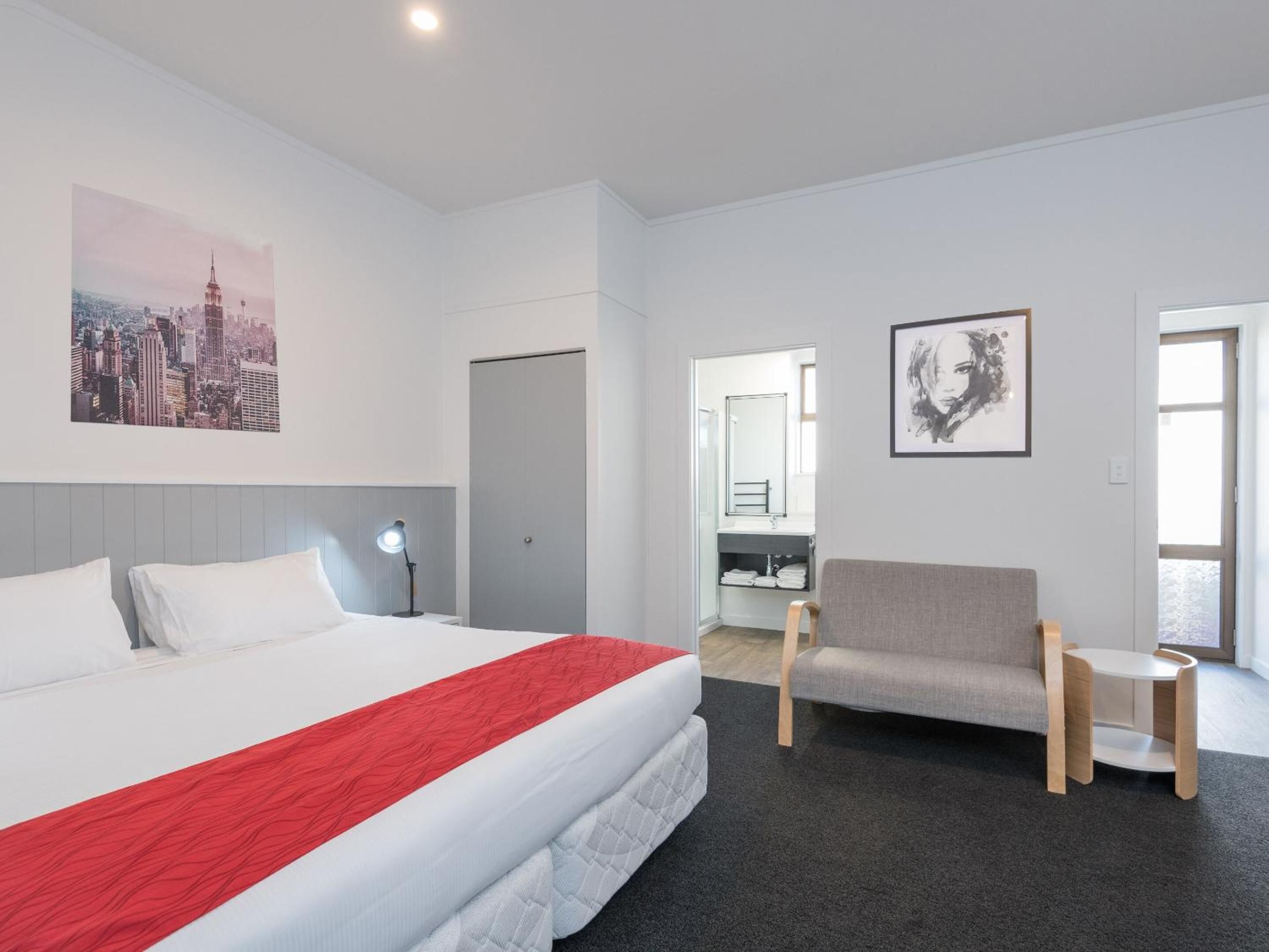 A hotel room with a large bed, a grey sofa, a small table, and a cityscape painting on the wall.