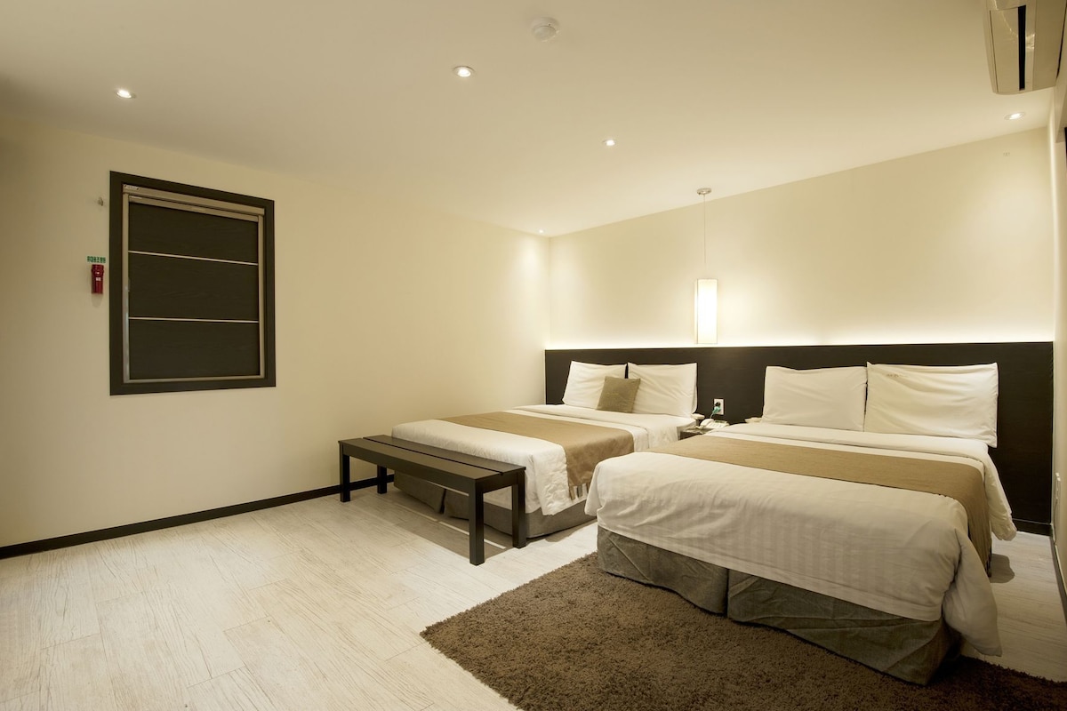 A hotel room with two beds, a bench, a wall-mounted light, and a ceiling vent.