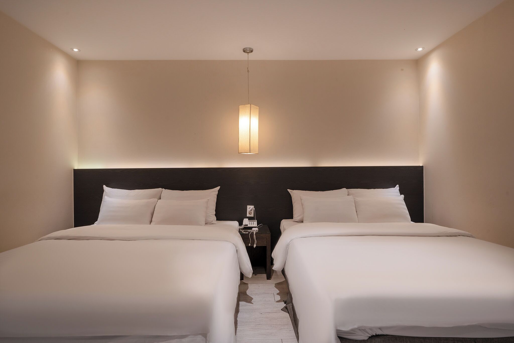 Una habitación de hotel con dos camas, un cabecero oscuro, una mesita de noche con lámpara e iluminación colgante.