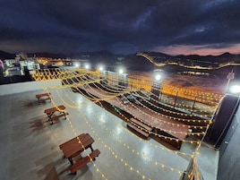An outdoor terrace with string lights, wooden benches, and tables, overlooking a cityscape at dusk.