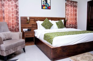 Um quarto com cabeceira de madeira, cama com roupa de cama branca e almofadas estampadas em verde, mesa de cabeceira com abajur e uma cadeira.