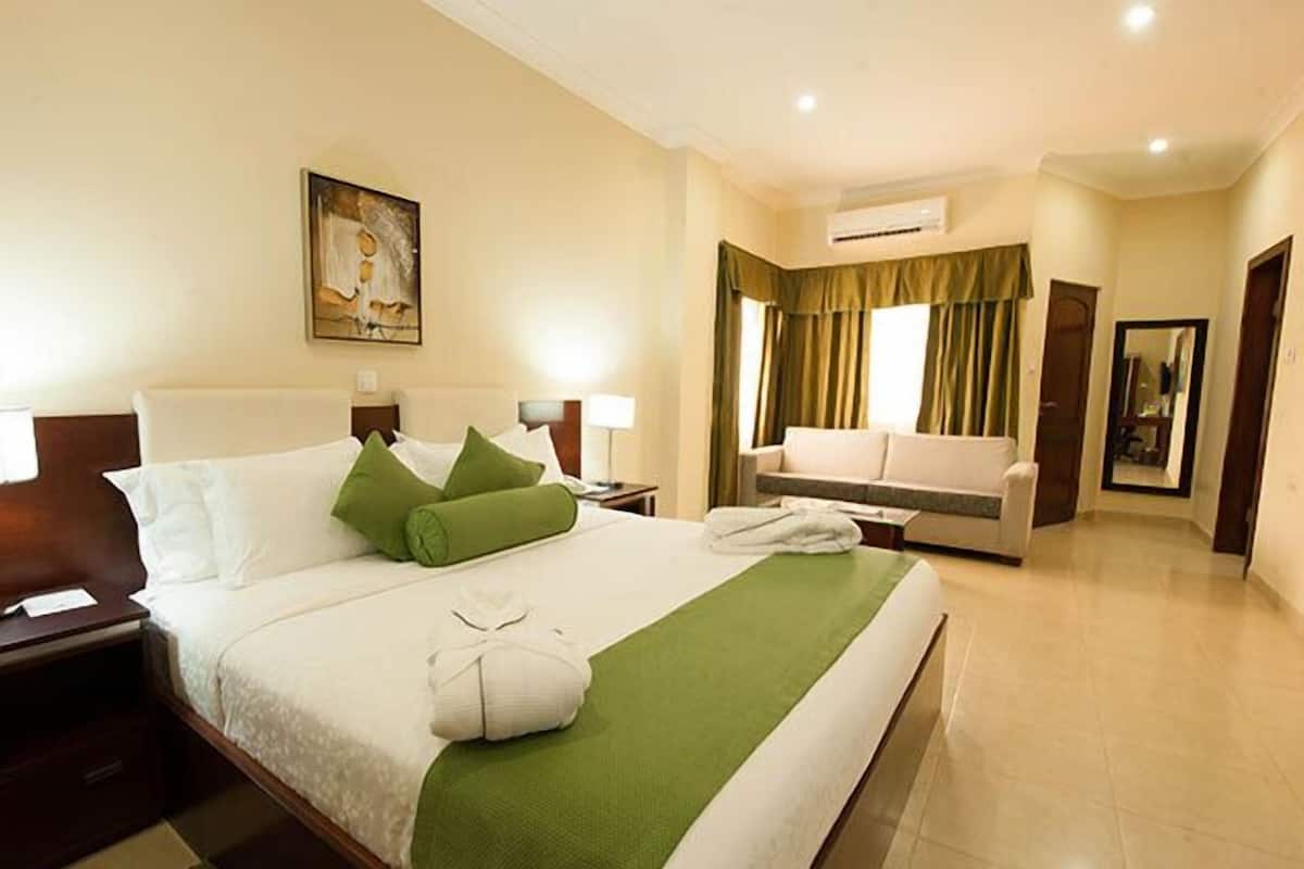 A hotel room with a large bed, a sofa, a painting on the wall, and an air conditioning unit.
