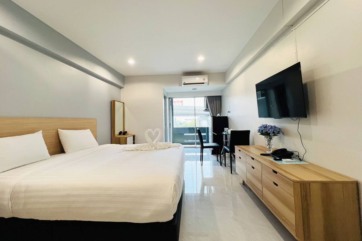A hotel room with a large bed, a wooden dresser, a desk with a chair, a flat-screen TV mounted on the wall, and a window with a view of the city.