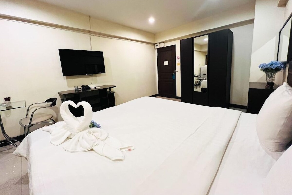 A hotel room with a bed, a flat-screen TV, a desk with a computer, a mirror, and a door leading to a bathroom.