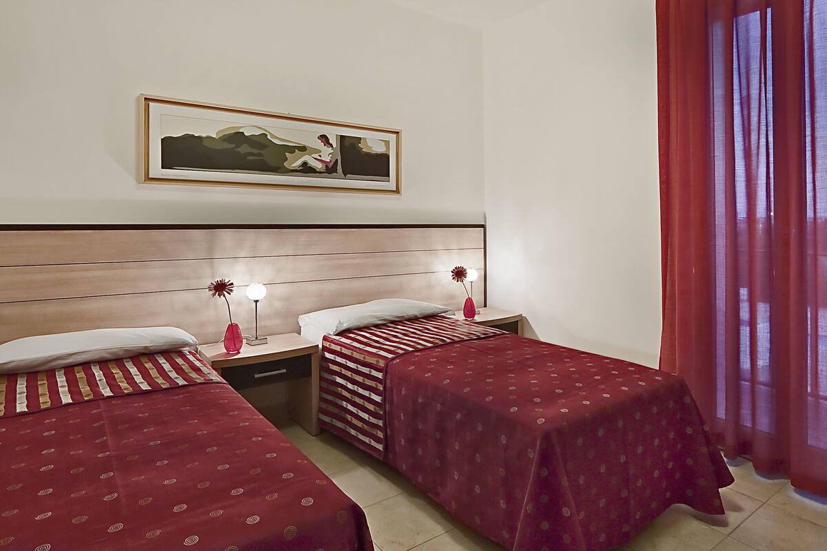 A hotel room with two beds, a nightstand with lamps, a framed picture on the wall, and a window with red curtains.