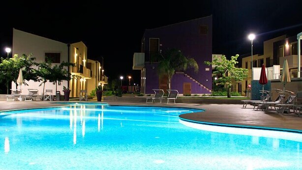 A nighttime swimming pool with lounge chairs and umbrellas, surrounded by buildings with balconies and outdoor seating. - Residence Blu ModicAmare (Modica)