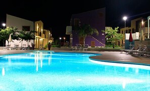 A nighttime swimming pool with lounge chairs and umbrellas, surrounded by buildings with balconies and outdoor seating. - Residence Blu ModicAmare (Modica)