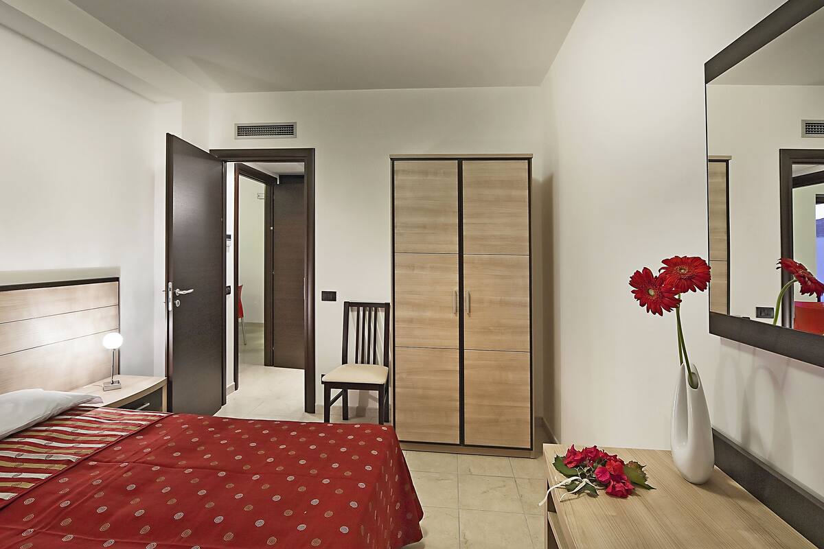 A bedroom with a bed, a nightstand, a wooden wardrobe, a chair, and a mirror.
