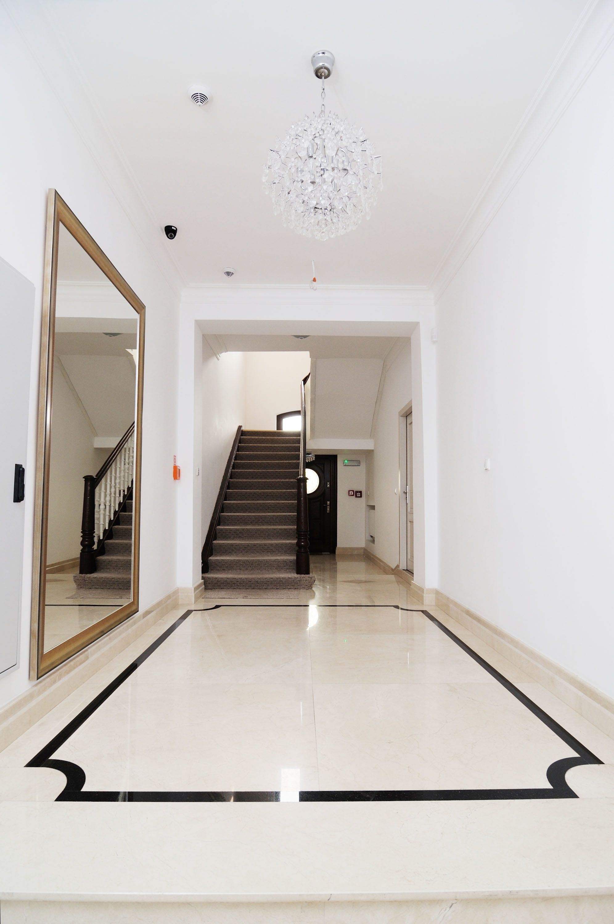Um corredor com piso de mármore, um espelho grande, um candelabro e uma escadaria que leva a outro cômodo.