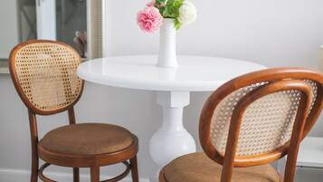 A dining area with two wooden chairs and a white table, a vase with flowers, and a white wall.