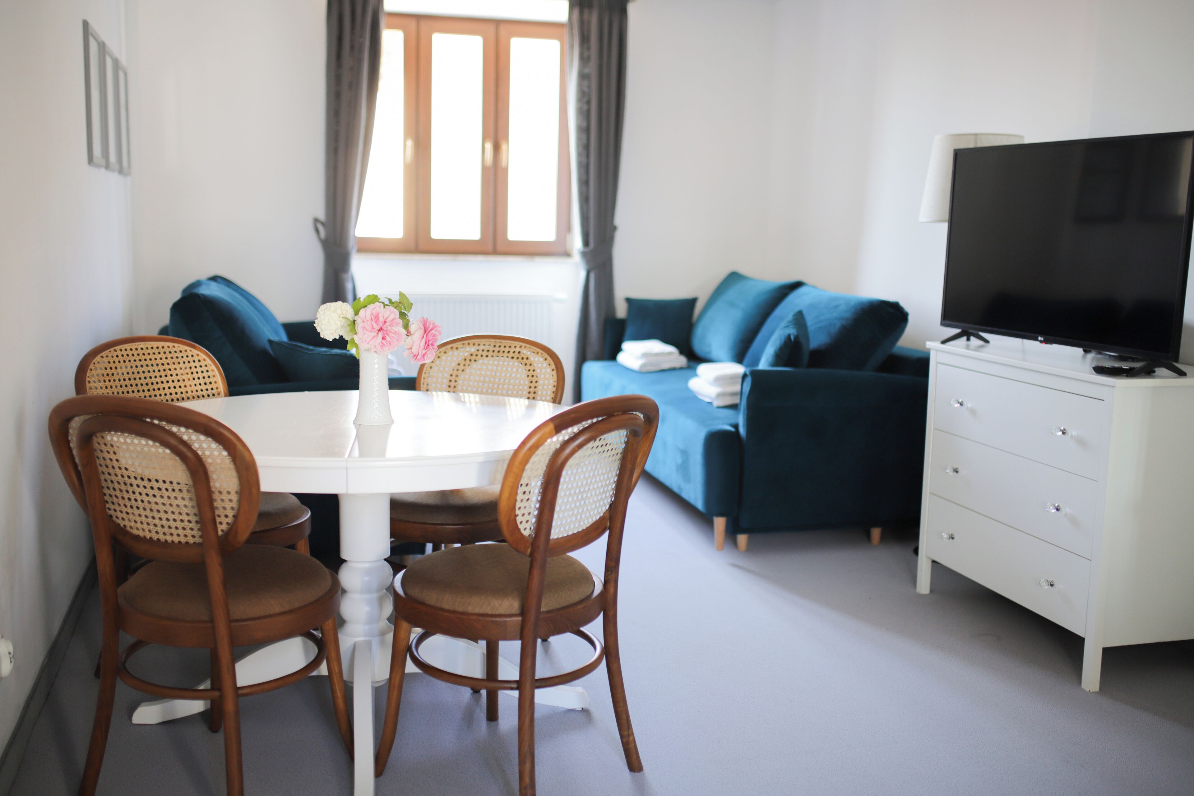 A modern living room with a dining table, two wicker chairs, a sofa, a TV, and a white dresser.