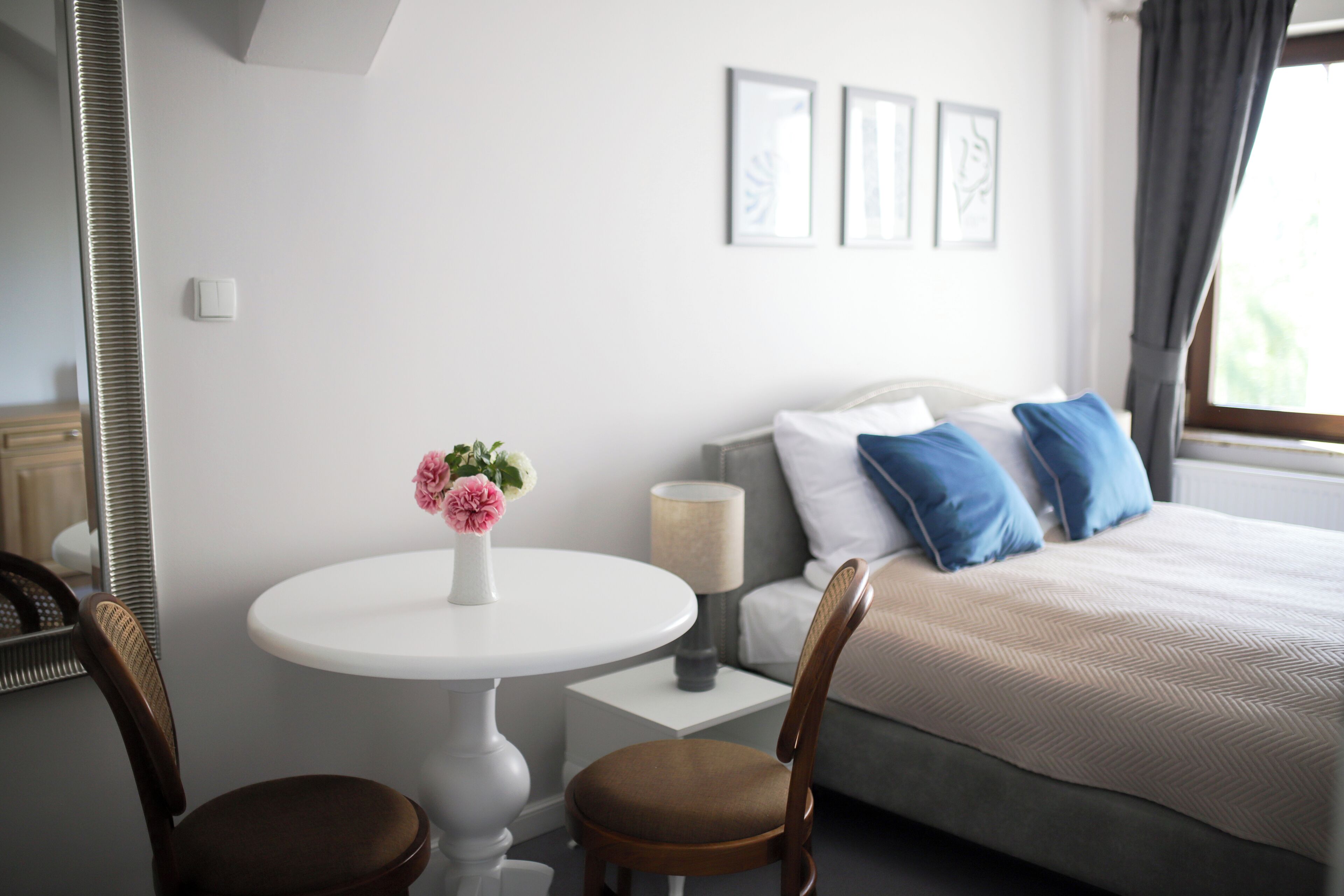 Una camera da letto con un letto, un comodino con una lampada, una sedia, un tavolino rotondo con un vaso di fiori e uno specchio.