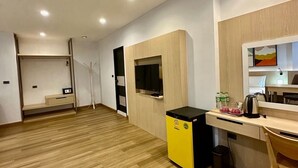 A modern hotel room with a wooden floor, a desk with a chair, a small kitchenette, a TV mounted on the wall, and a bed in the background. - Sakura Sky Residence (Bangkok)
