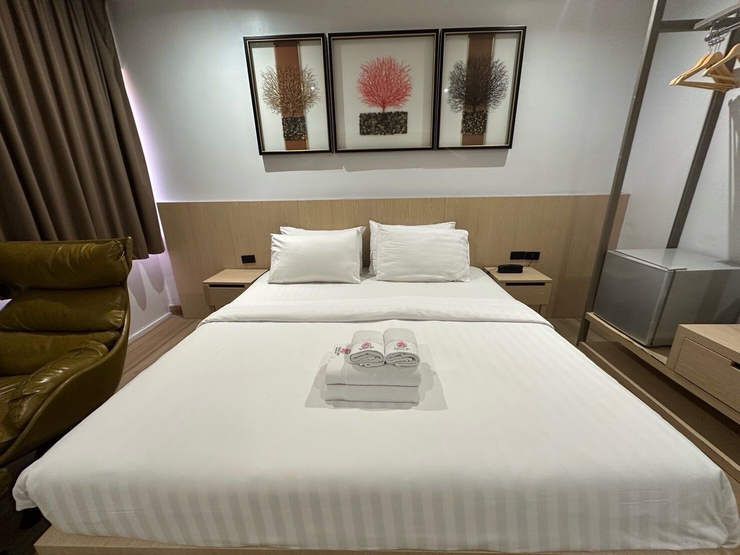 Une chambre d’hôtel avec un lit soigneusement fait, deux tables de chevet, un fauteuil en cuir marron et trois œuvres d’art encadrées accrochées au mur.