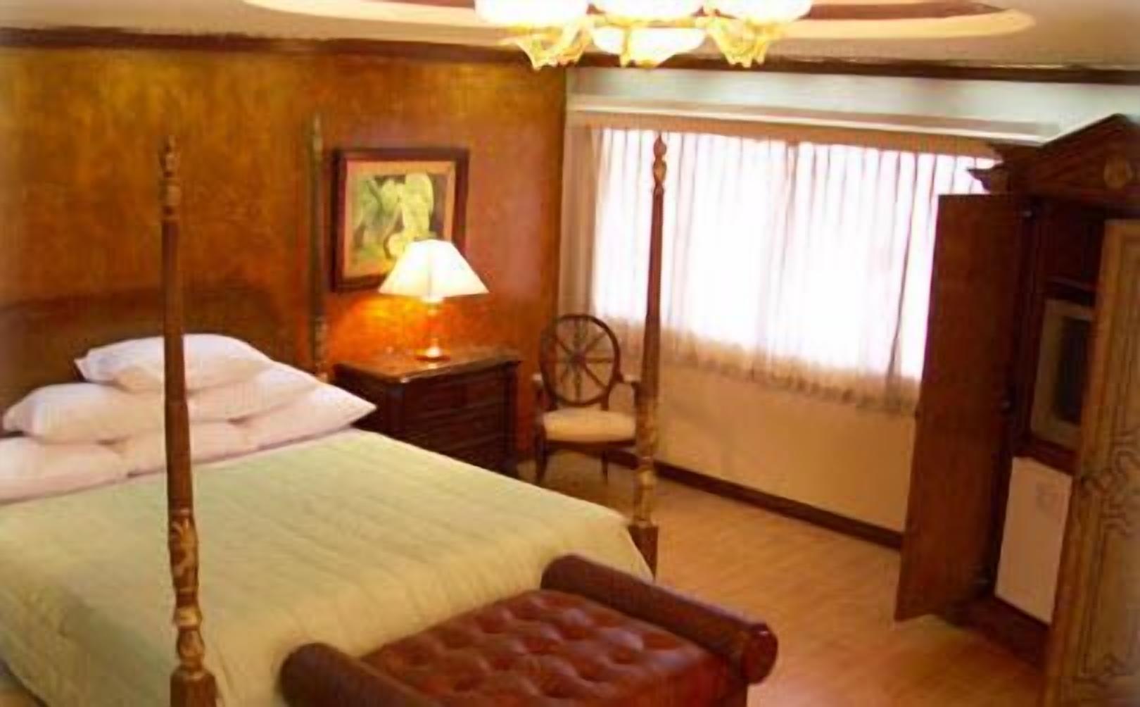 A four-poster bed with a wooden headboard, a nightstand with a lamp, a chair, and a window with curtains.