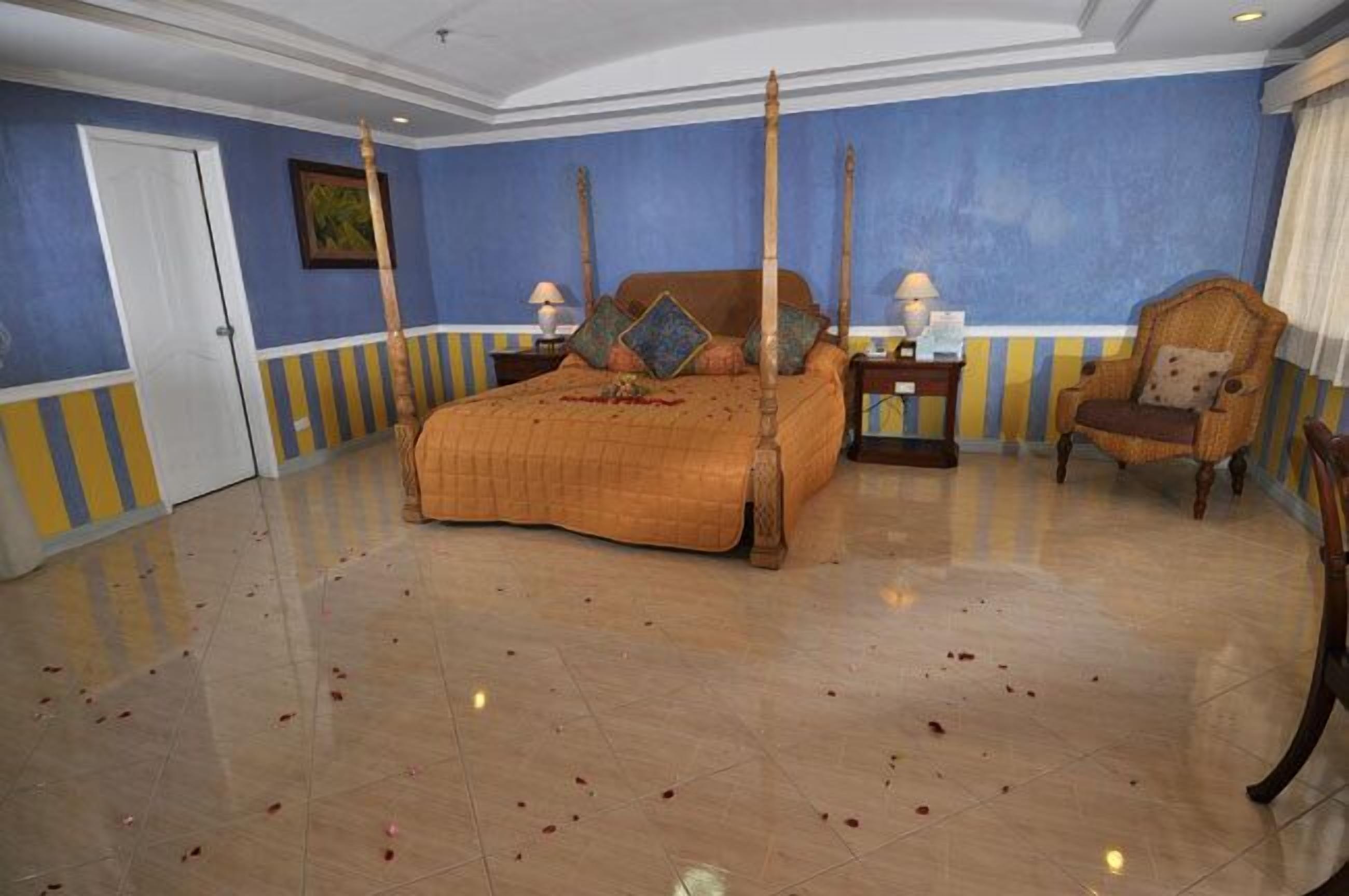 A four-poster bed with a wooden frame, a large bedspread, and multiple pillows. There are two bedside tables with lamps, a chair, and a door in the room.