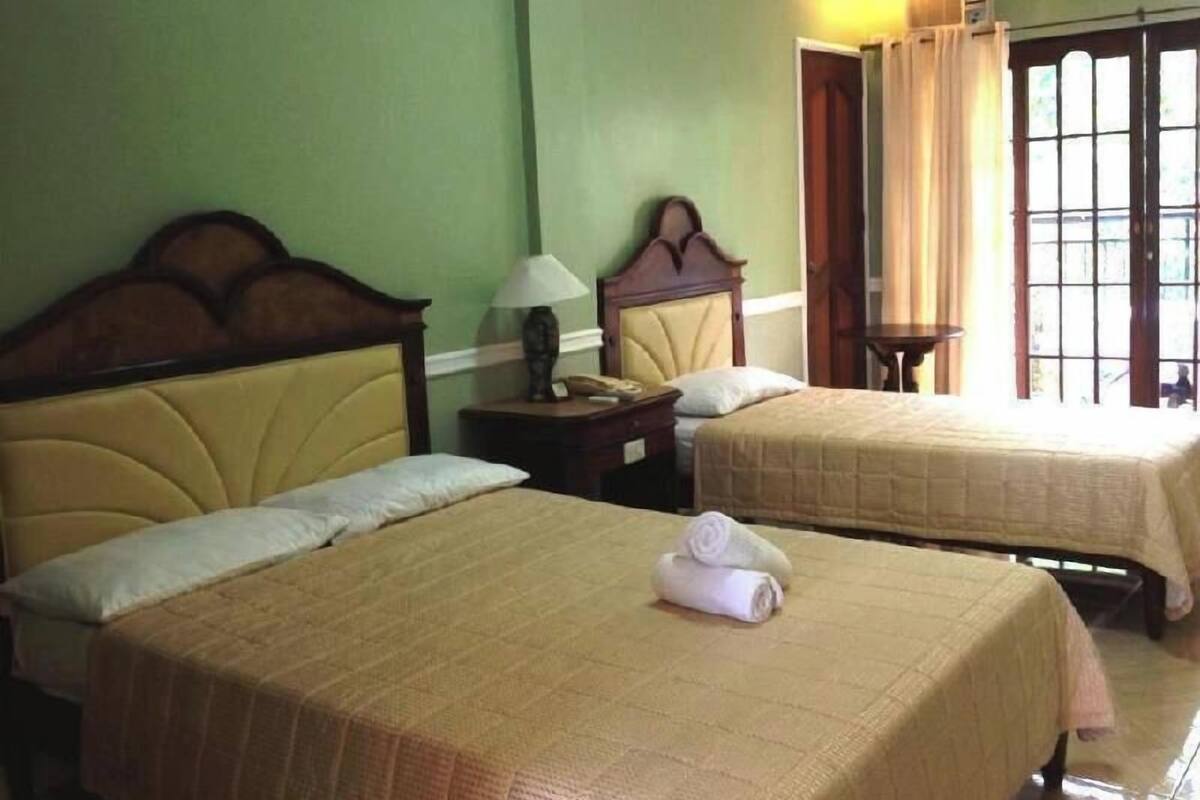 A hotel room with two beds, a nightstand with a lamp, a window with curtains, and an air conditioning unit.
