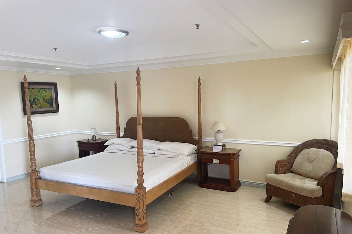 A four-poster bed with a wooden headboard, a nightstand with a lamp, a wicker armchair, and a small table with a lamp.