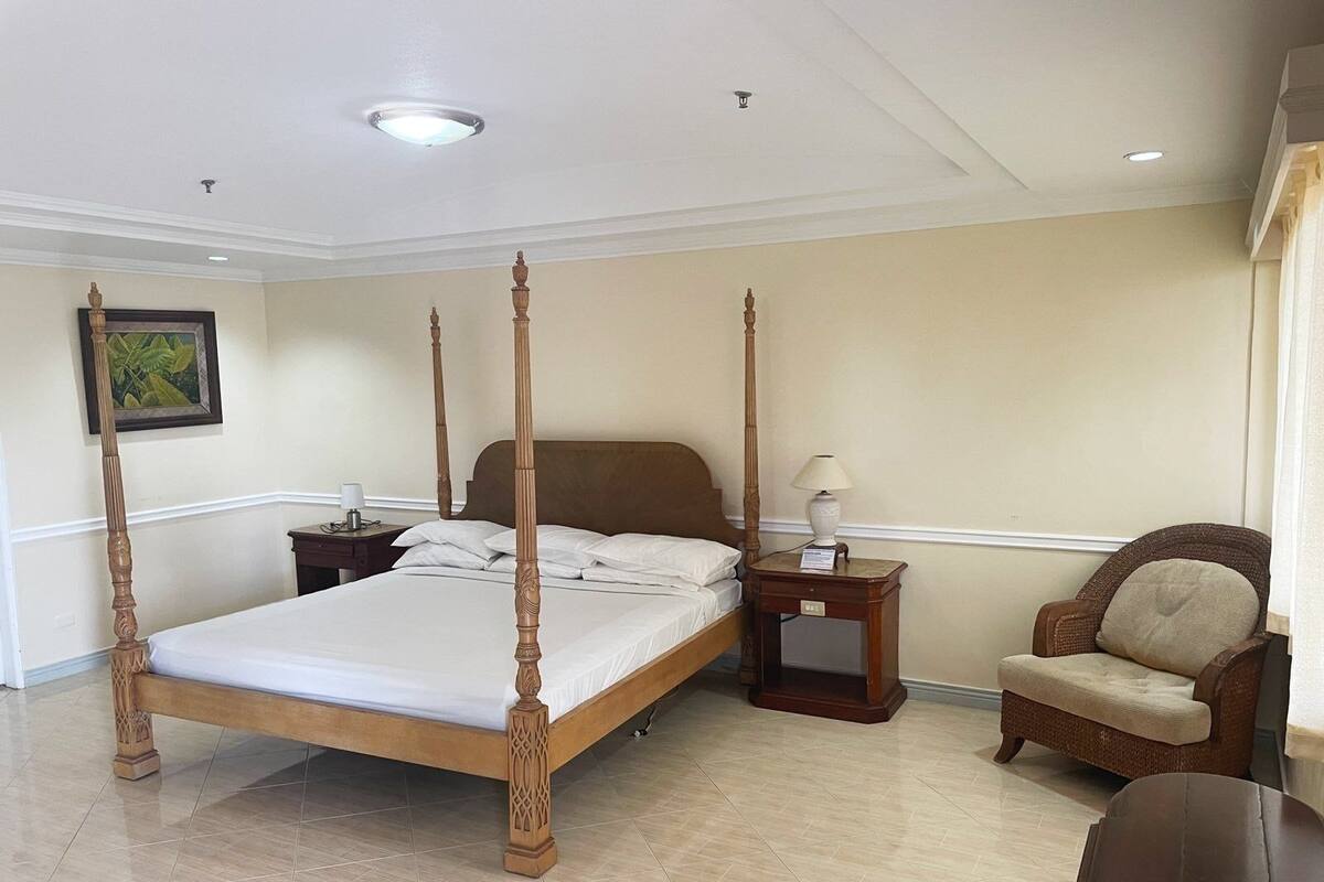 A four-poster bed with a wooden headboard, a nightstand with a lamp, a wicker armchair, and a small table with a lamp.