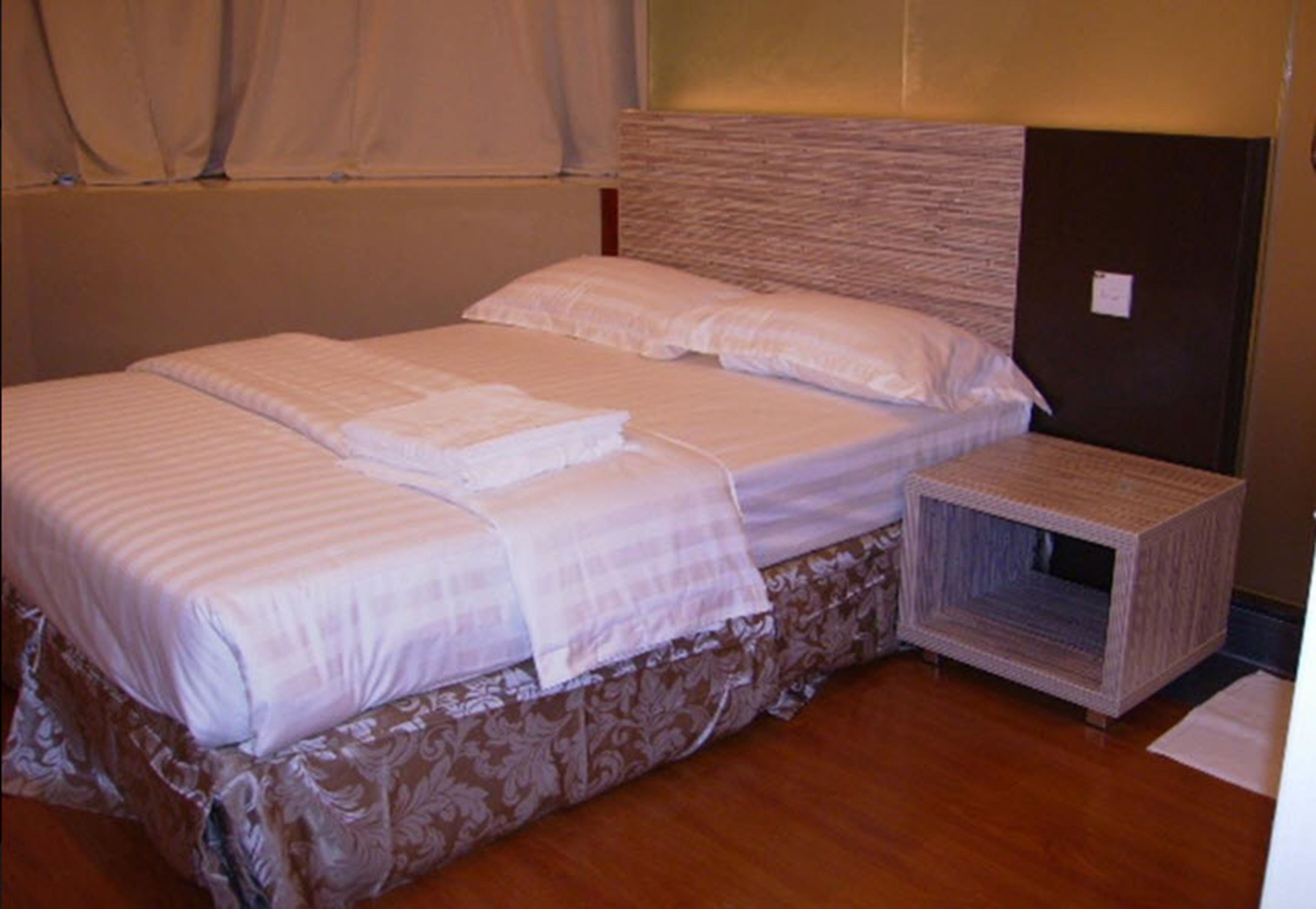 A neatly made bed with a patterned bedspread and pillows, a small bedside table with a woven cover, and a headboard with a textured design.