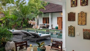 A poolside area with lounge chairs, a dining table, and a view of a house with a tiled roof. - Room & Vespa 3 (Seminyak)