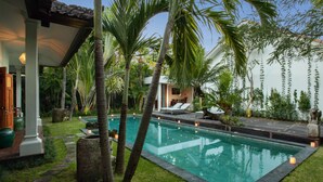 A swimming pool with a stone border, surrounded by palm trees and a covered seating area. - Room & Vespa 3 (Seminyak)