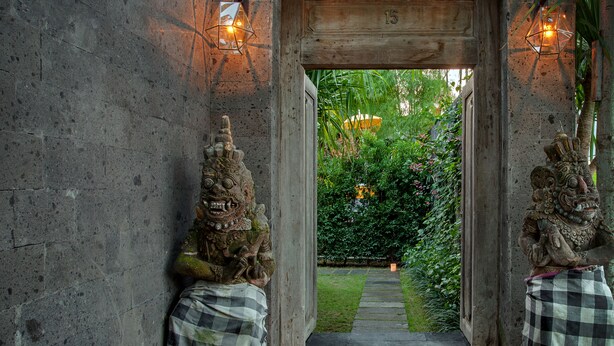 A stone pathway leading to a wooden archway with two decorative statues on either side, each draped with a checkered cloth. - Room & Vespa 3 (Seminyak)
