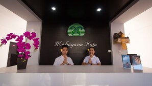 A reception desk with a logo and name "Mochayana Kuta" on the wall. There are two individuals standing behind the desk, and a potted plant with purple flowers to the left. - RaBaSTa Mahayana Kuta (Kuta)