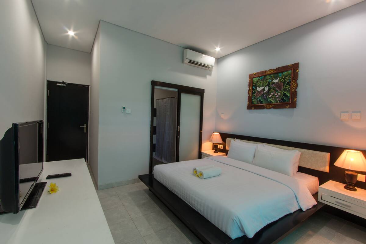 A hotel room with a bed, bedside tables, a desk with a computer, and an air conditioning unit.