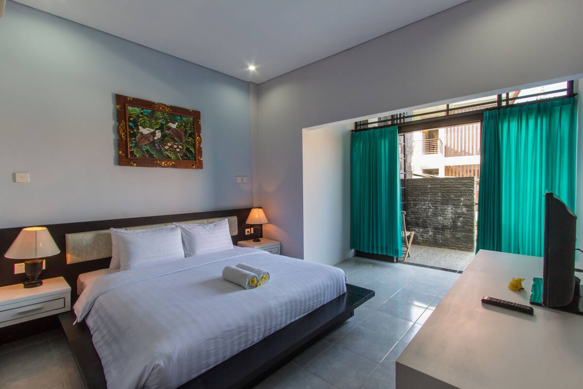 A modern hotel room with a large bed, bedside lamps, a painting on the wall, and a view through a door with curtains.