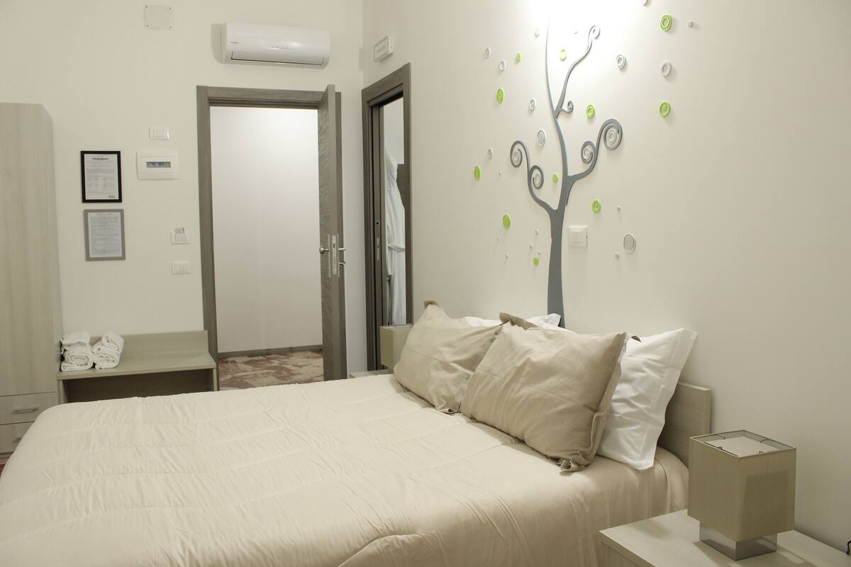 A hotel room with a bed, a nightstand, a wall-mounted tree decoration, and a door leading to another room.