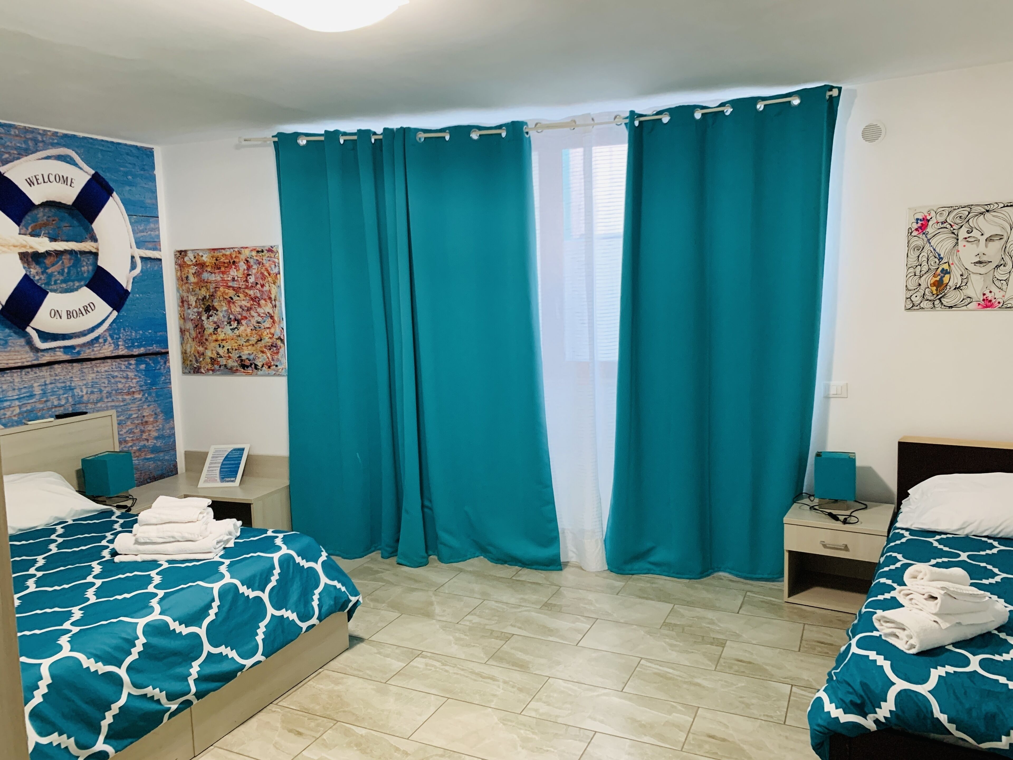 Habitación de hotel con dos camas, cortinas color turquesa, decoración con un salvalavidas, un cuadro abstracto colorido y suelo de baldosas.