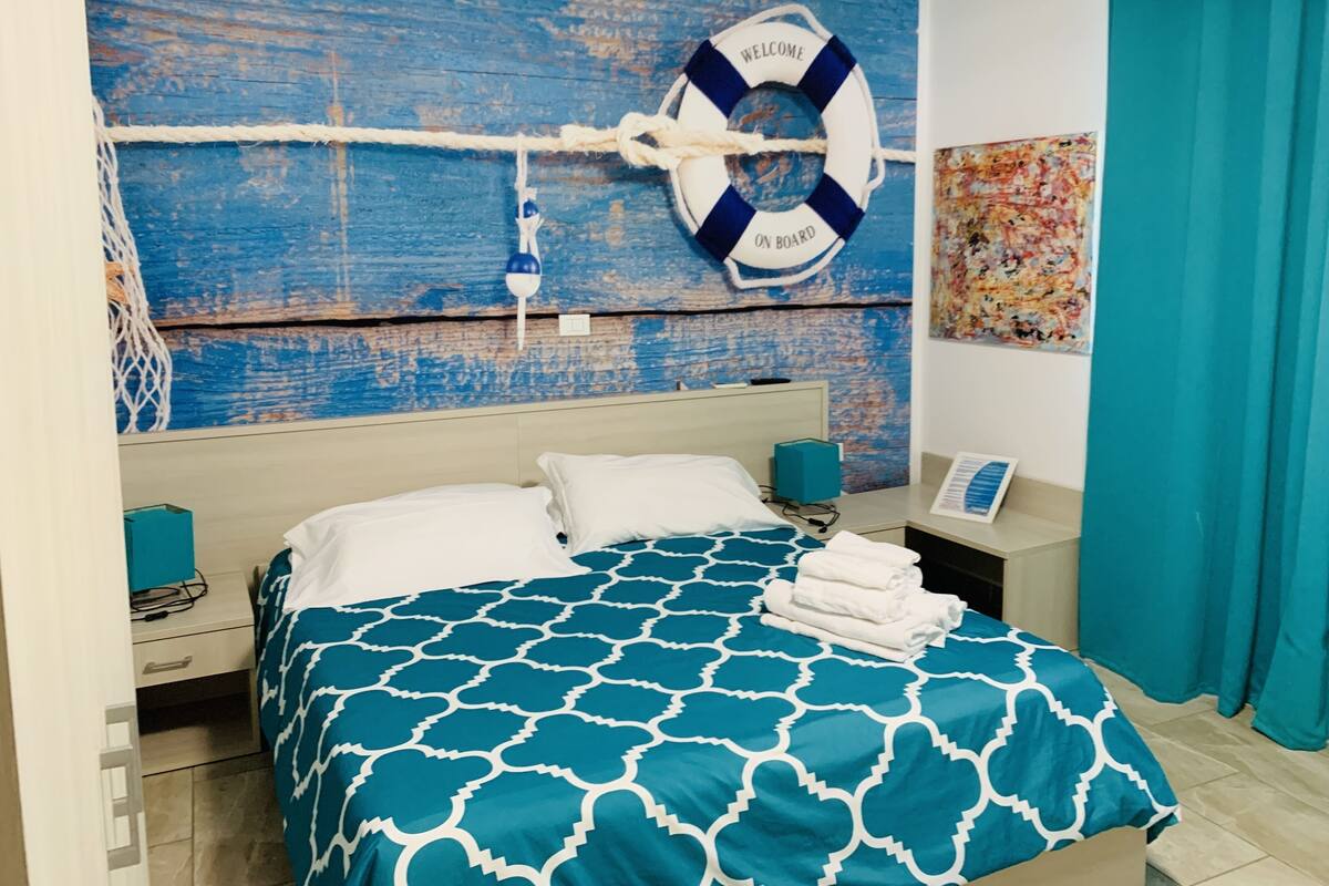 A neatly made bed with a blue and white patterned bedspread, a life preserver decoration on the wall, and a colorful abstract painting.