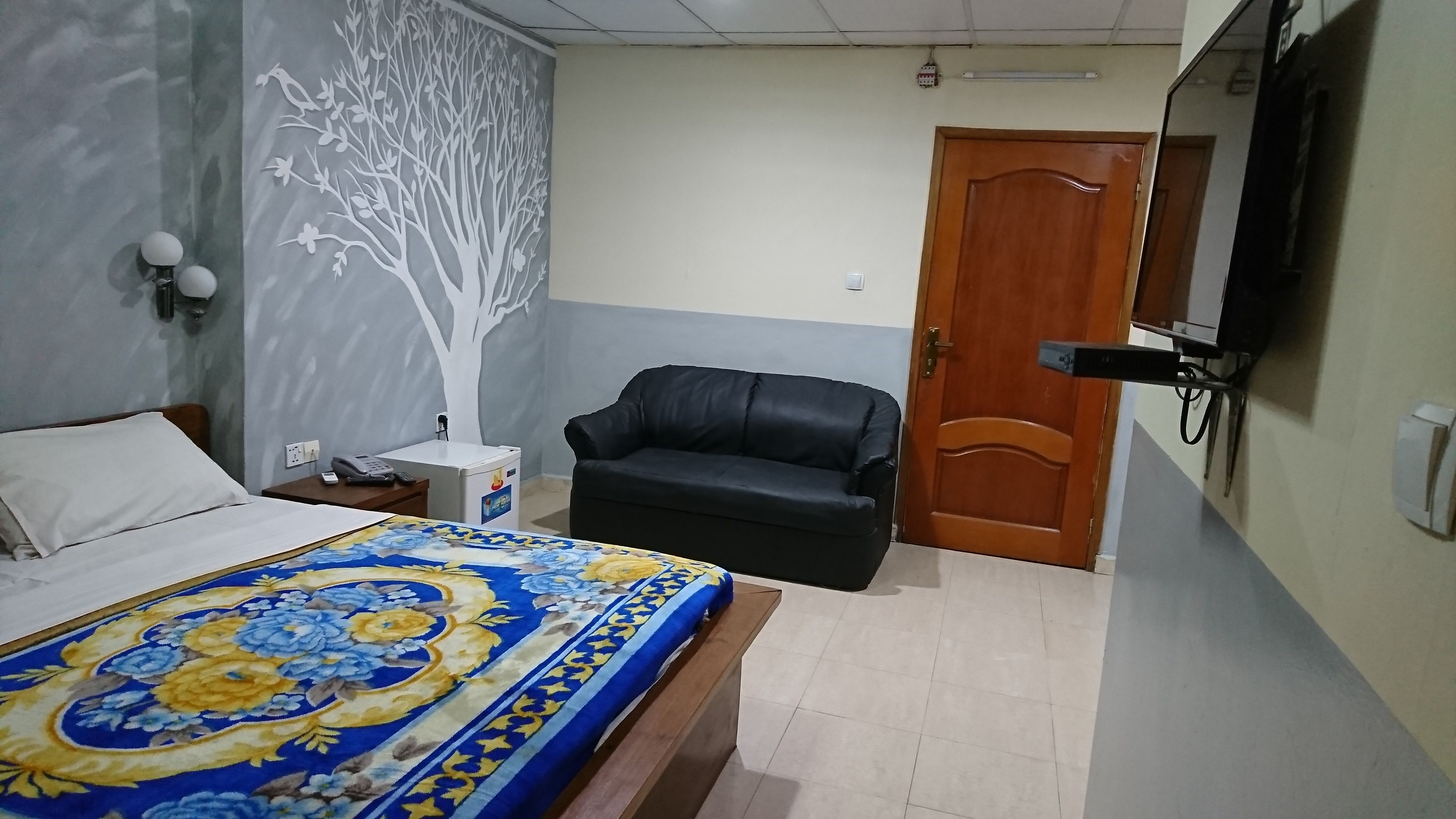 A bedroom with a bed, a black leather chair, a wooden door, and a wall with a tree mural.