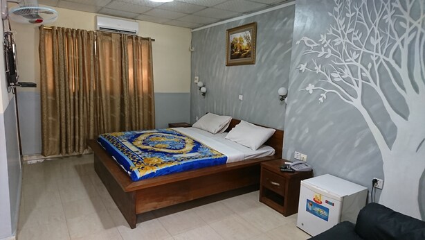 A bedroom with a bed, bedside tables, a heater, an air conditioner, a refrigerator, and a wall with a tree design. - Hotel Pour Vous (Kinshasa)