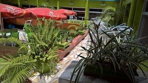 An outdoor seating area with green chairs and tables, red umbrellas, and potted plants. - Hotel Pour Vous (Kinshasa)