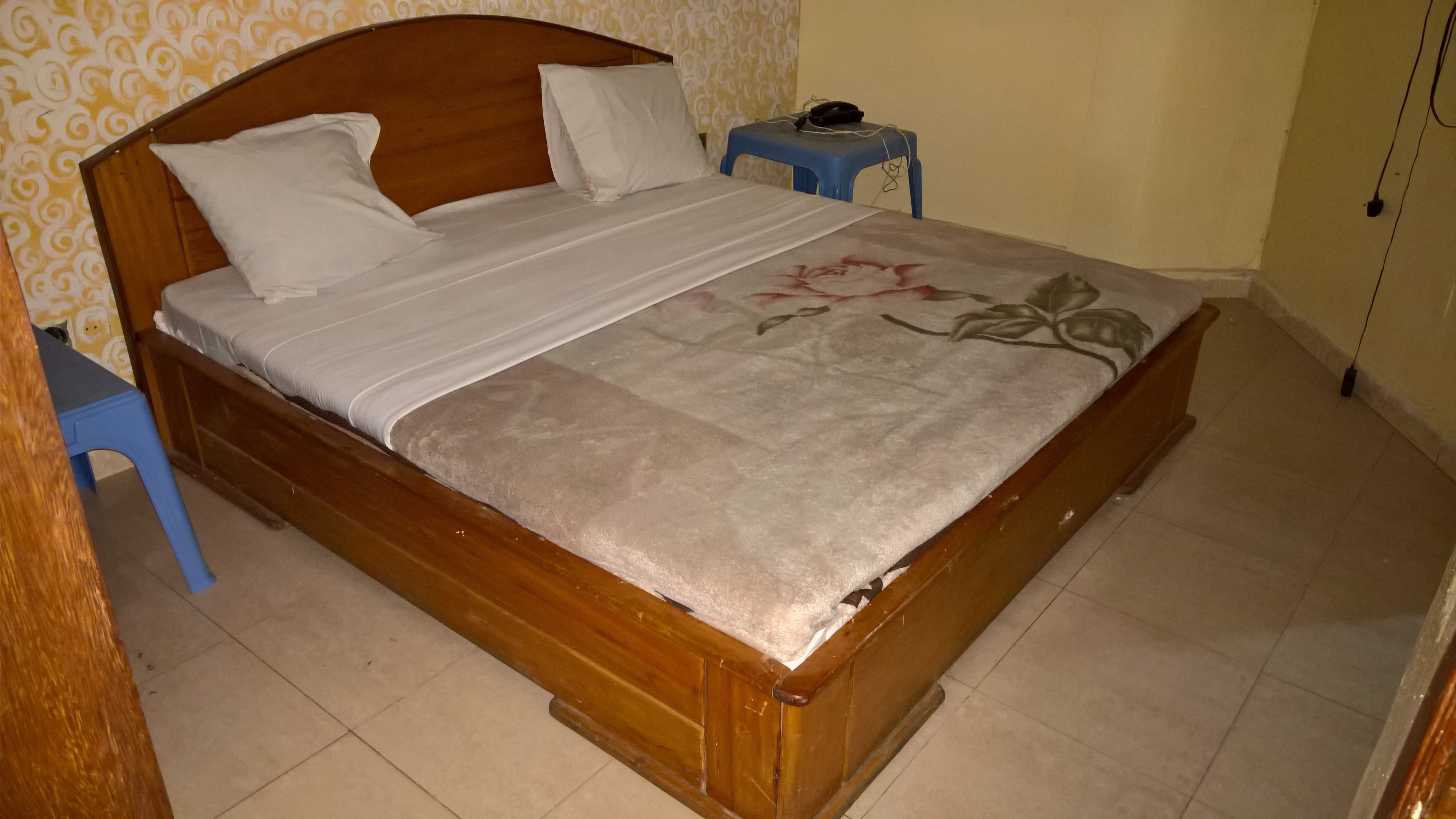 Una cama de madera con una colcha estampada de flores, dos almohadas, una mesita de noche azul pequeña con un teléfono, y una pared con papel tapiz estampado.