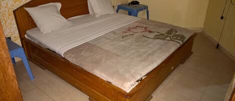 Una cama de madera con una colcha estampada de flores, dos almohadas, una mesita de noche azul pequeña con un teléfono, y una pared con papel tapiz estampado.