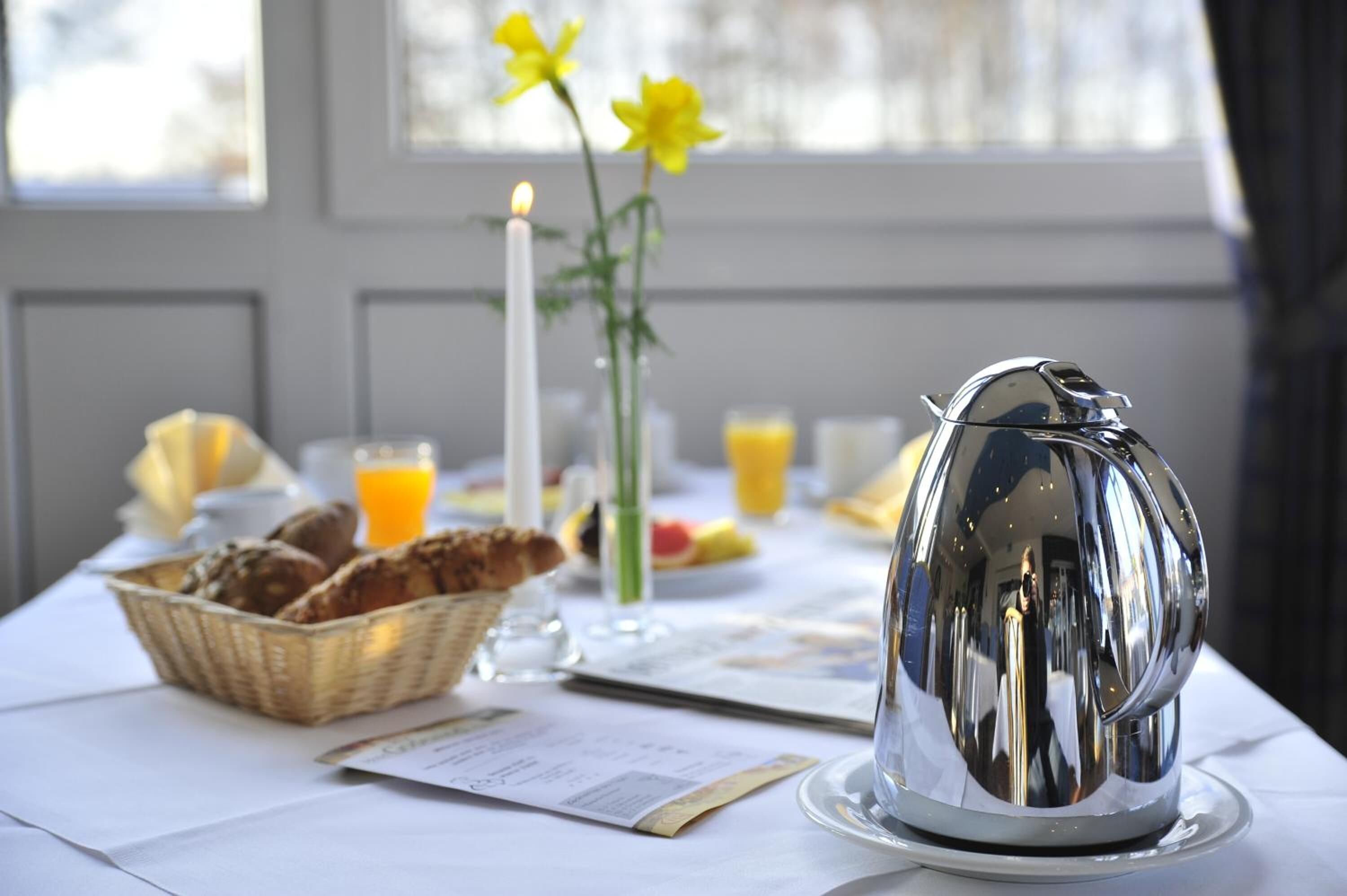 Une table mise pour le petit-déjeuner avec une théière en argent, un panier de pain, un vase rempli de fleurs jaunes, une bougie allumée et un verre de jus d’orange.