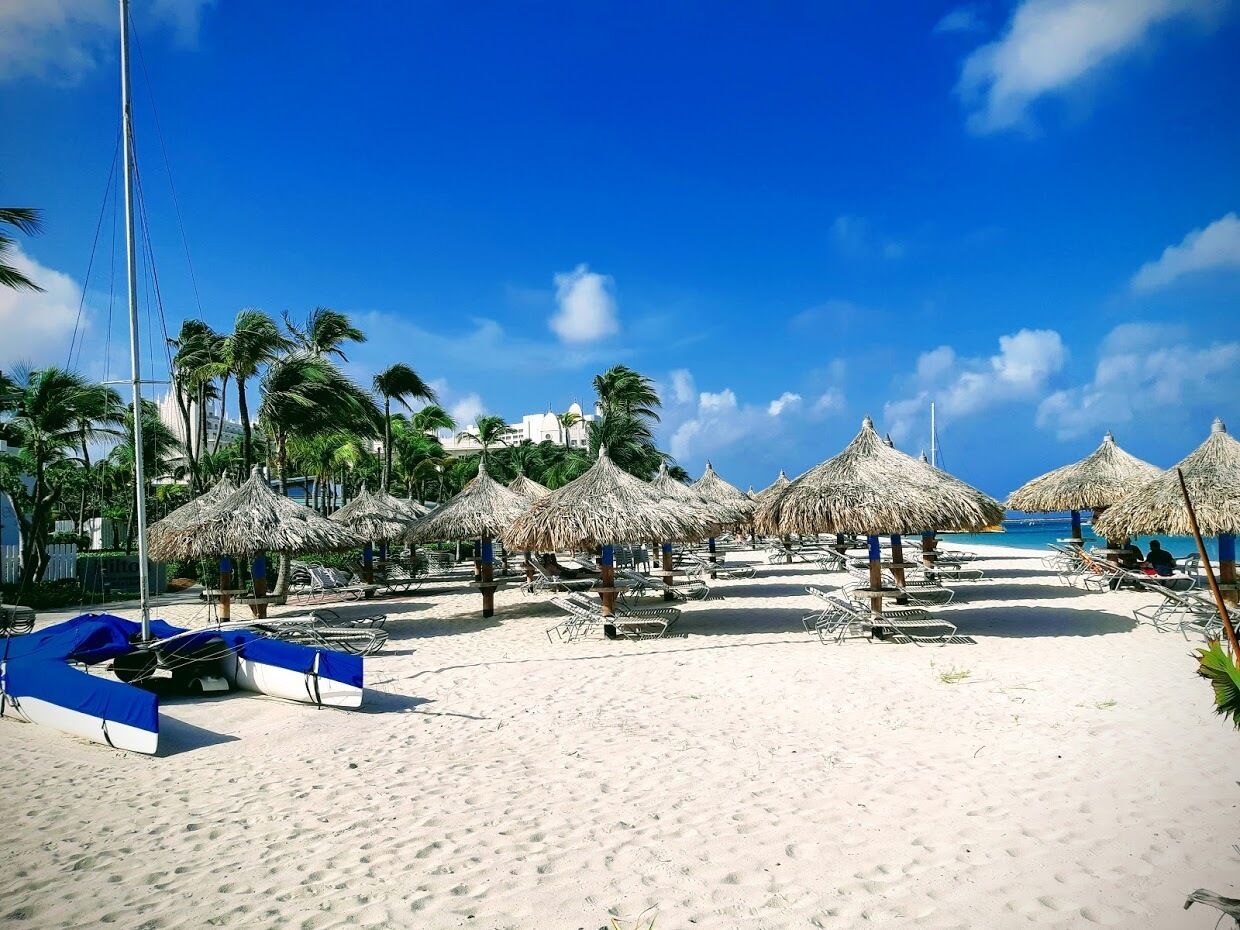 A beach with thatched umbrellas, lounge chairs, and a sailboat.