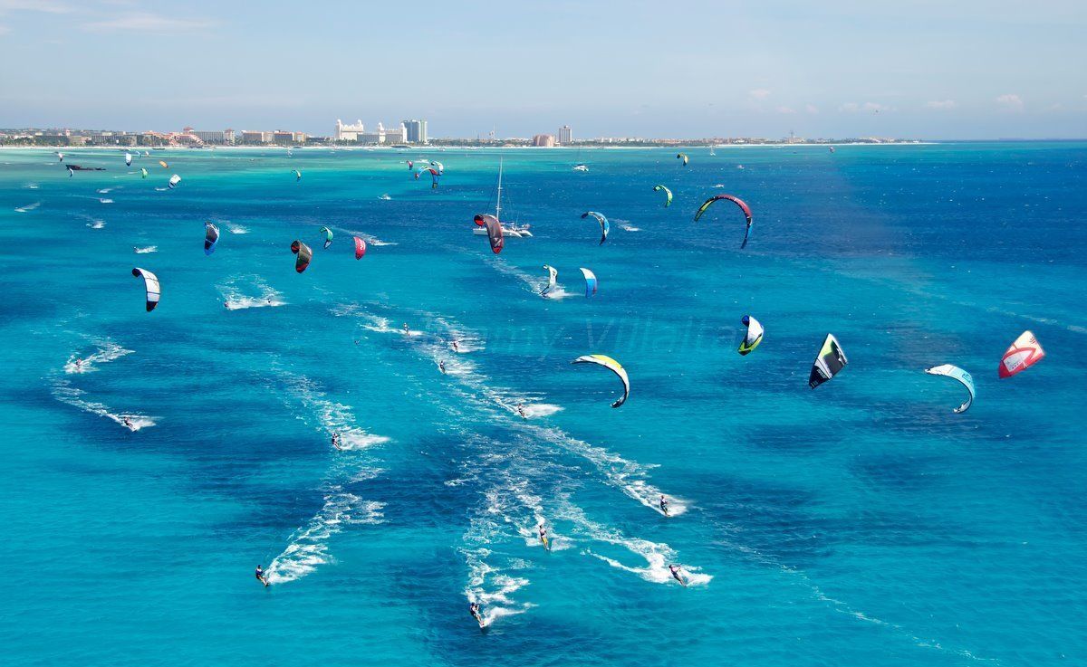 Zona costera con varios kitesurfistas en el agua, mar azul cristalino y edificios a lo lejos.