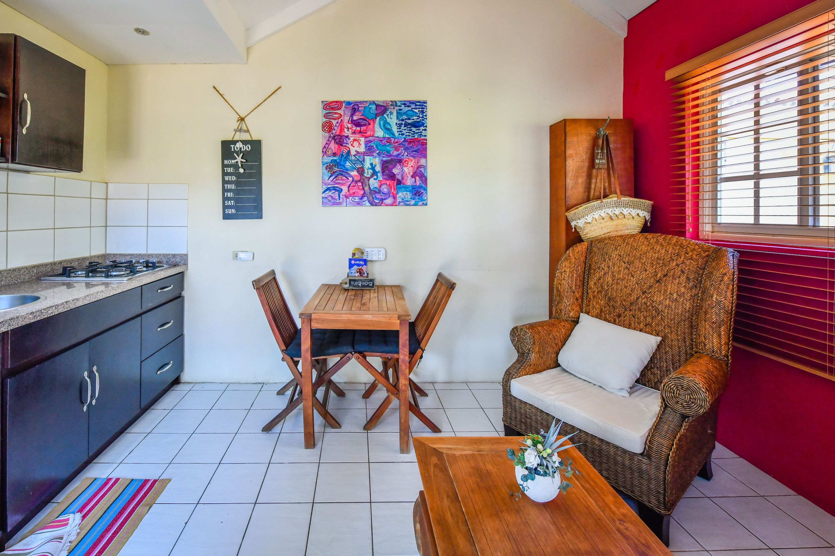 A compact kitchen with a gas stove, dark cabinets, and a tiled floor. There is a wooden dining table with chairs and a wicker sofa with a cushion. A colorful abstract painting hangs on the wall, and a basket is placed on the sofa.