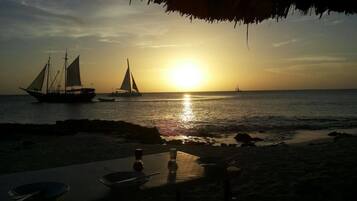 A beach scene with a table set for dining, overlooking the sea with sailboats.