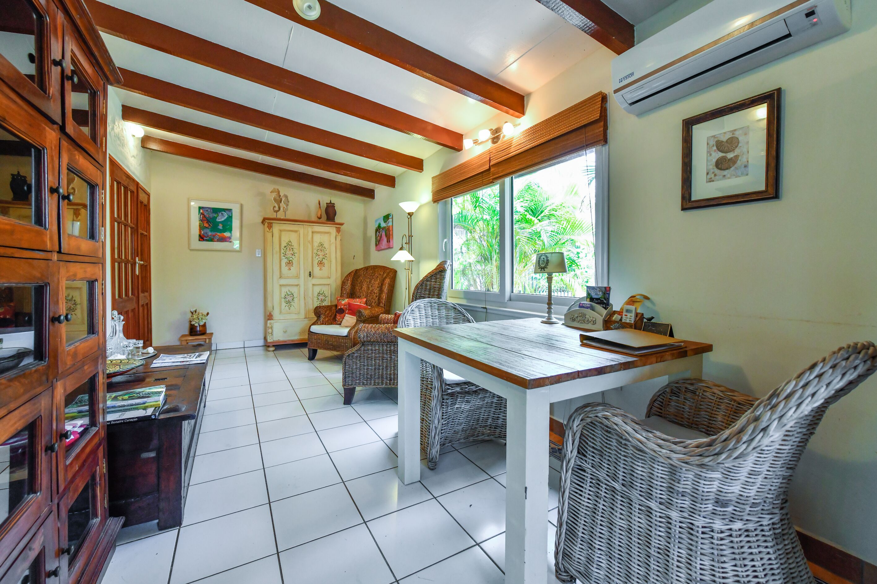 A dining area with a wooden table and wicker chairs, a cabinet with glass doors, and a window with a view of greenery.