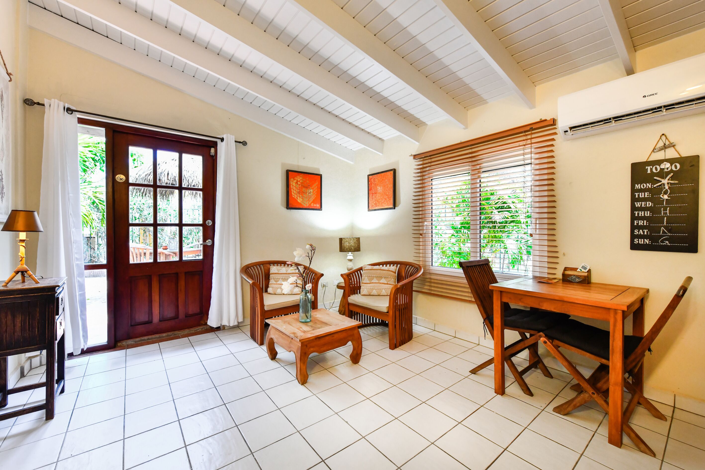 A room with a wooden table, two wicker chairs, a wooden coffee table, a lamp, and a calendar on the wall.