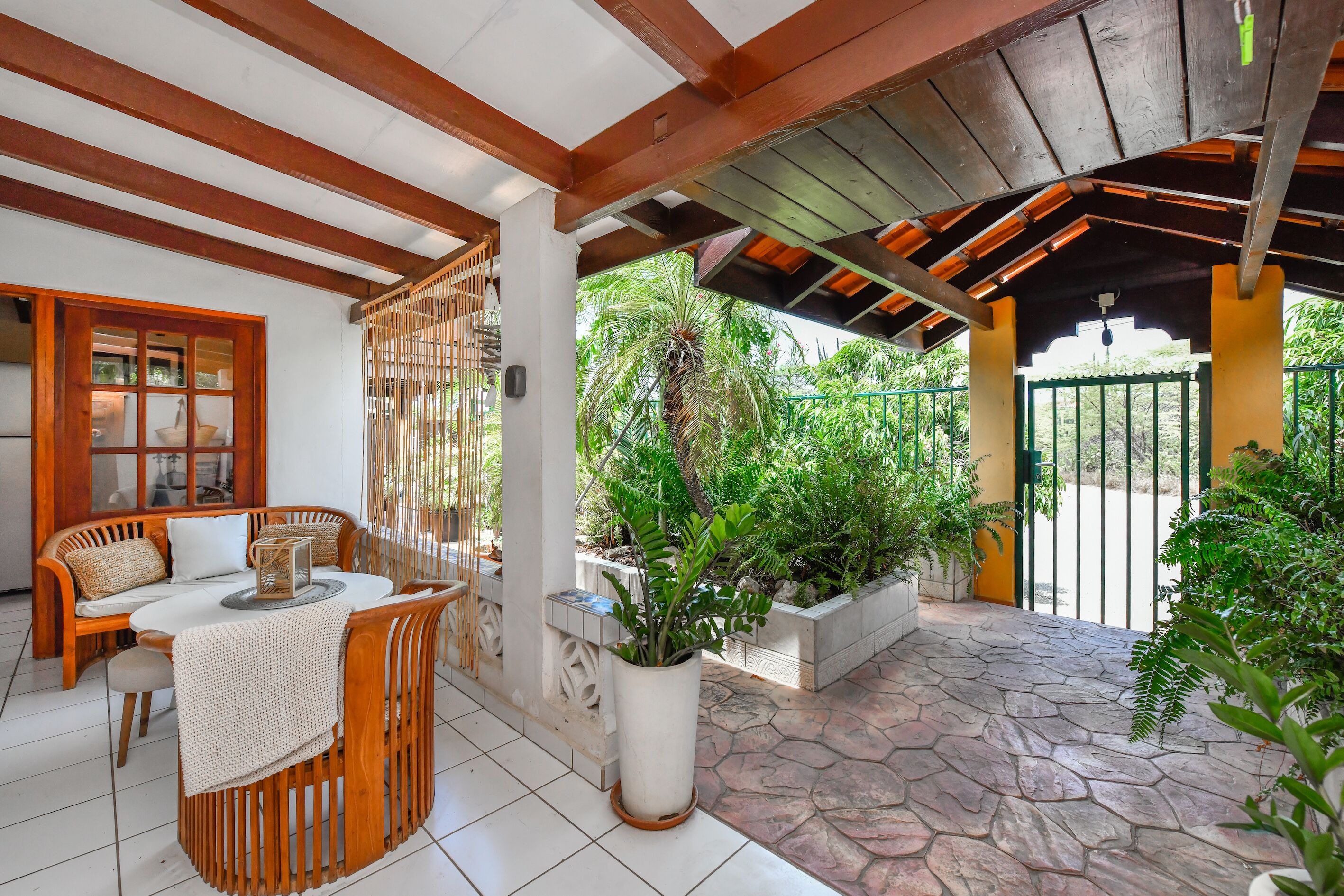 Un patio techado con un banco y mesa de madera, plantas en macetas y vistas del follaje circundante.